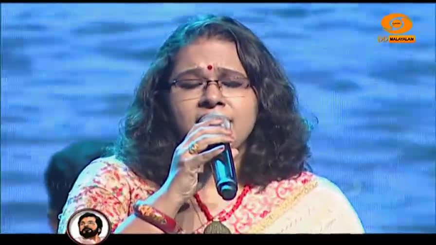 A woman with glasses sings into a microphone, her eyes closed in concentration. The DD Malayalam logo appears in the upper right corner.