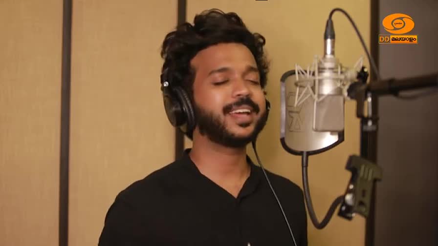 A man with headphones on, eyes closed, sings into a microphone in a recording studio. The DD Malayalam logo is visible in the corner.