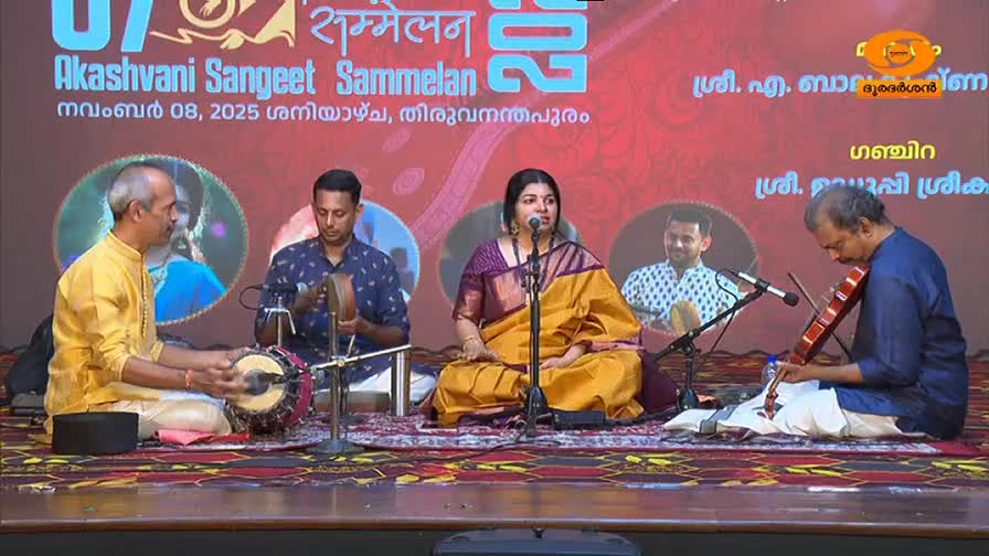 A woman in a bright yellow and purple sari sings into a microphone, her hands resting on her lap. To her left, a man in a yellow kurta plays a traditional Indian drum, while another man to her right plays the violin. The backdrop behind them displays text in Malayalam and English, announcing "Akashvani Sangeet Sammelan" in Thiruvananthapuram.