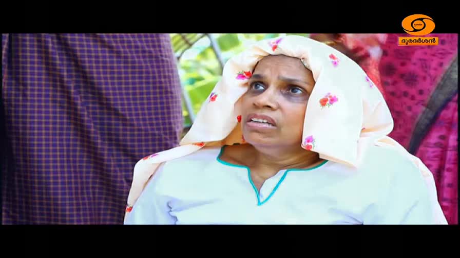 A woman with a worried expression is looking upwards, her head covered by a light pink scarf patterned with small flowers. Beside her, a person in a dark purple checkered lungi stands partially visible.