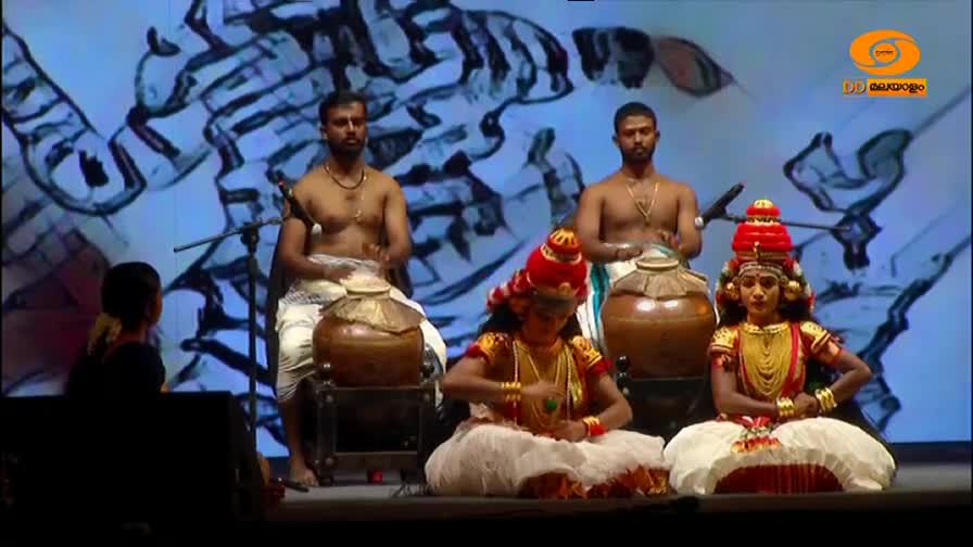Two men in white dhotis beat on large clay pots, their bare chests gleaming under the lights. In front of them, two dancers in vibrant costumes, one with an elaborate red headdress, sit poised.