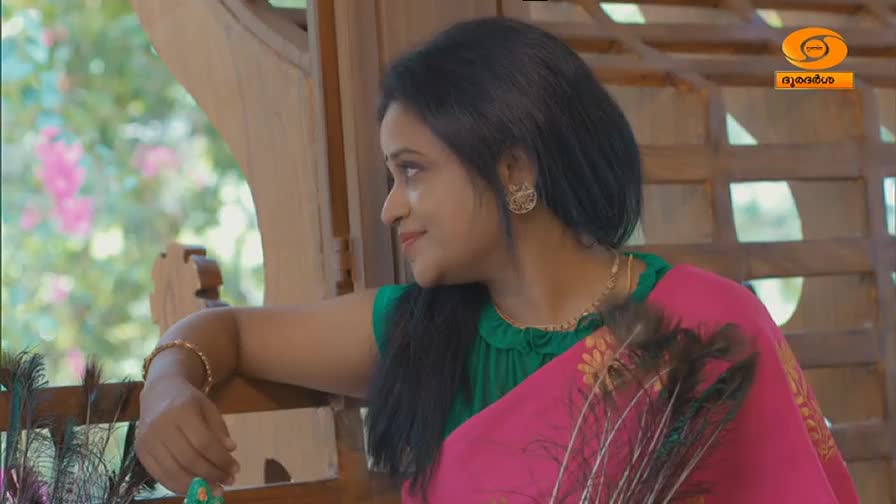 A woman in a bright pink sari with a green blouse looks out a window, a gentle smile on her face. The DD Malayalam logo is visible in the corner, hinting at a scene from a television program.