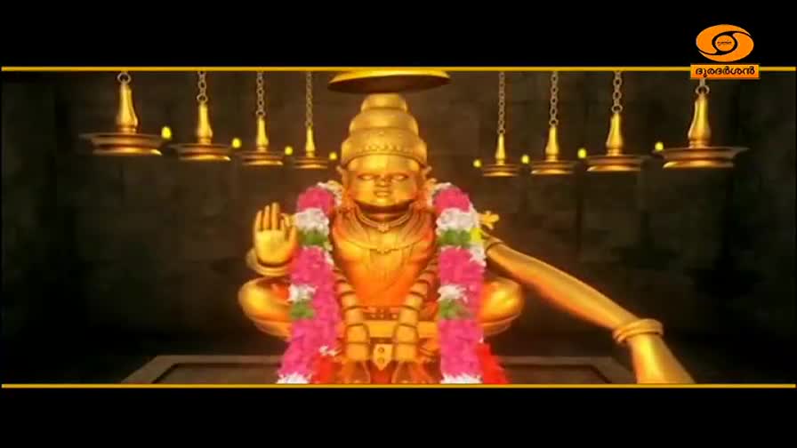 A golden idol of a deity sits cross-legged, draped in garlands. Above, brass oil lamps hang from chains, their flames casting a warm glow. This is a scene from DD Malayalam, a channel broadcasting from India.