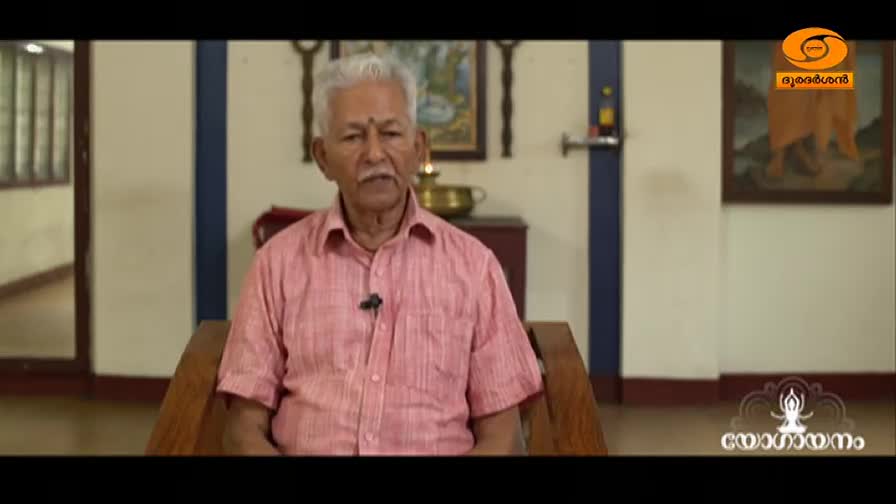 An elderly man with white hair and a mustache speaks directly to the camera. He wears a pink striped shirt and sits on a wooden chair in a room with traditional Indian decor. The DD Malayalam logo appears in the corner.