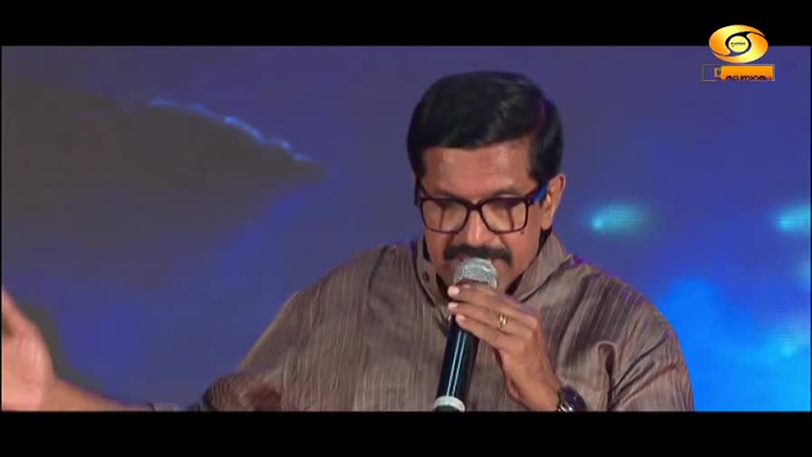 A man with glasses and a mustache speaks into a microphone on a stage. The DD Malayalam logo is visible in the corner.