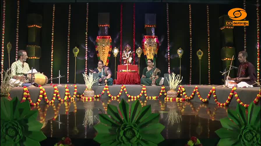 A man plays the mridangam to his left, while another man bows a stringed instrument to his right. Two women sit between them, facing forward as a statue of a deity presides over the stage. The DD Malayalam logo is visible in the upper right corner.