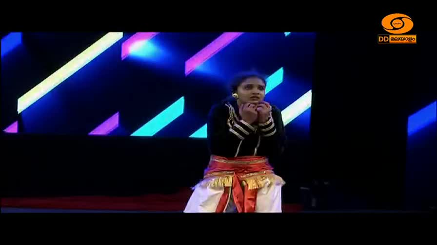 A young woman stands on stage, her hands clasped near her face, her expression intense. Behind her, bright diagonal lines of light flash across a dark backdrop, a typical sight during a DD Malayalam broadcast.