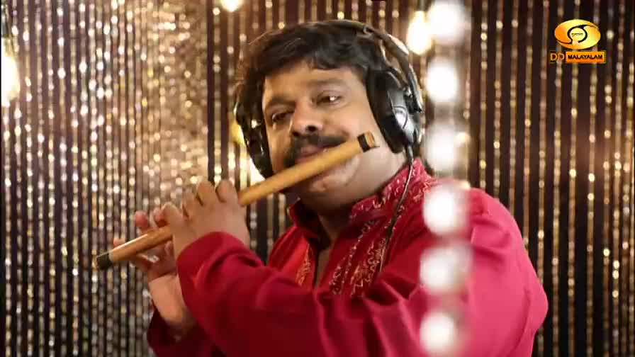 A man in a red shirt and headphones plays a bamboo flute, his fingers moving over the holes. The DD Malayalam logo appears in the corner, set against a backdrop of shimmering gold curtains.