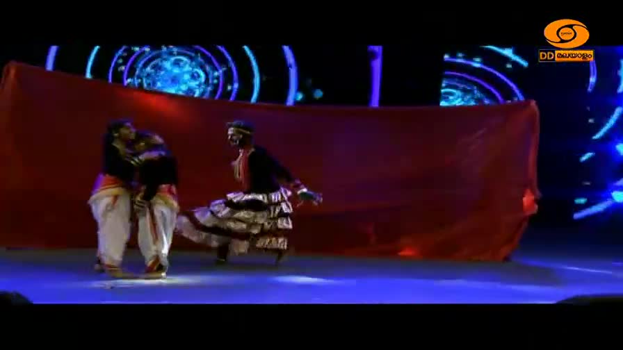 Two dancers in traditional Indian attire move across the stage, one in white dhoti and the other in a layered skirt. A large red cloth hangs behind them, obscuring part of the backdrop.