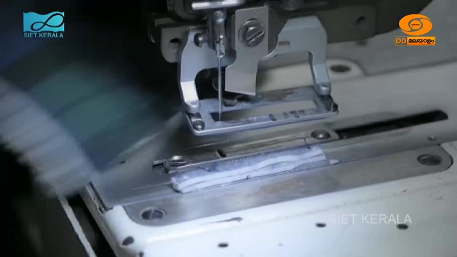 The needle of the industrial sewing machine plunges down, stitching a strip of fabric. A roll of white material feeds into the machine from the left, its edge blurred by rapid movement.
