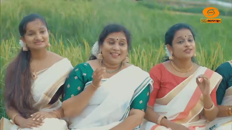 Three women in white saris with gold borders sit in front of a green field, their hands gesturing as they sing. The DD Malayalam logo is visible in the upper right corner.