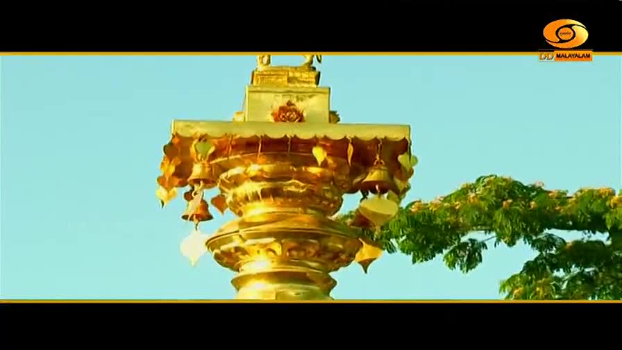 A golden structure, adorned with small bells, stands against a clear blue sky. Lush green foliage drapes over the side of the frame, framing the ornate temple element.