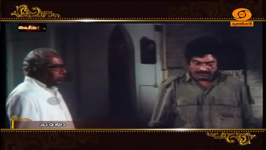 An older man in a white shirt stands to the left, while a man in a khaki uniform with a stern expression faces forward. This scene is part of a program on DD Malayalam, indicated by the channel's logo.