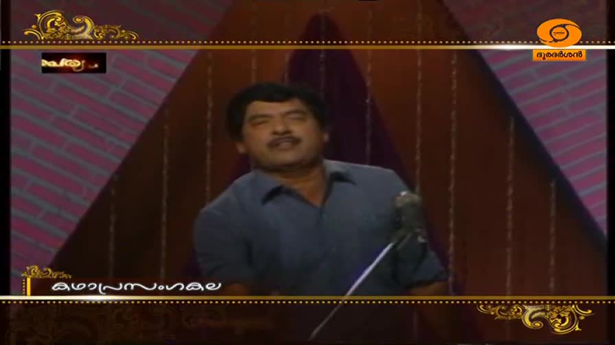 A man in a blue shirt stands at a microphone, his head tilted back as if in mid-sentence. The DD Malayalam logo appears in the corner of the screen.