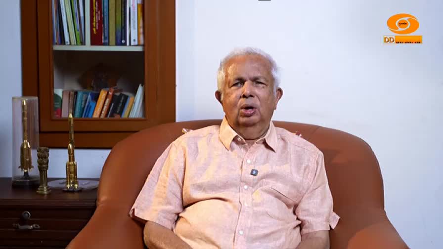 An older gentleman sits in a plush armchair, speaking into a microphone. Behind him, a bookshelf is filled with volumes, and the DD Malayalam logo is visible in the corner.