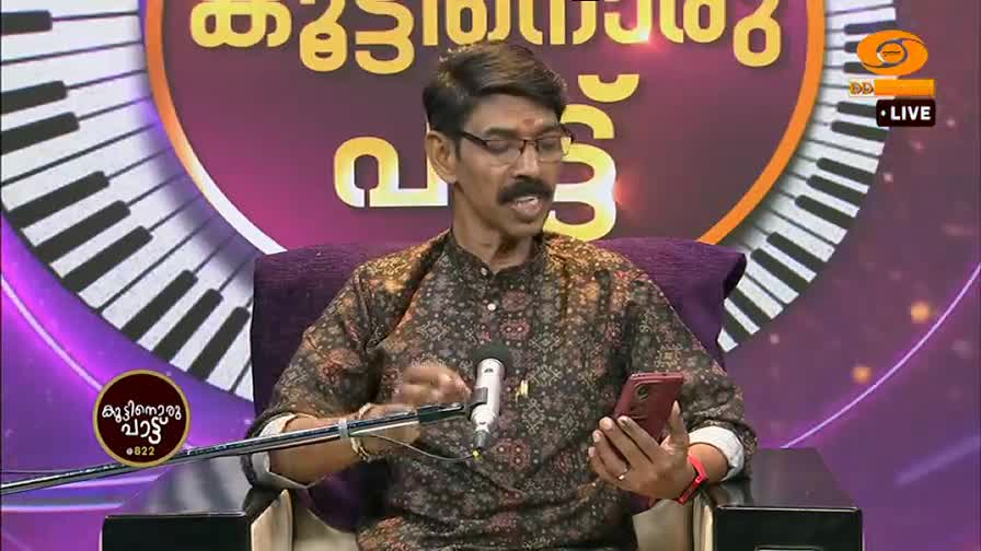 A man with a mustache and glasses is speaking into a microphone, holding a red phone in his other hand. The DD Malayalam logo is visible behind him, next to a graphic of piano keys.
