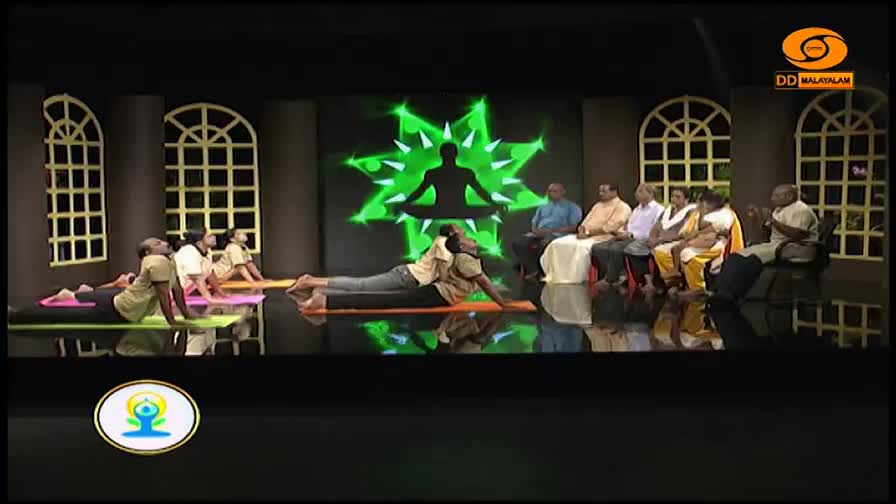 A group of people are performing yoga poses on mats. A man in the foreground is arching his back, his chest lifted off the floor. A panel of seated individuals watches them from the side.