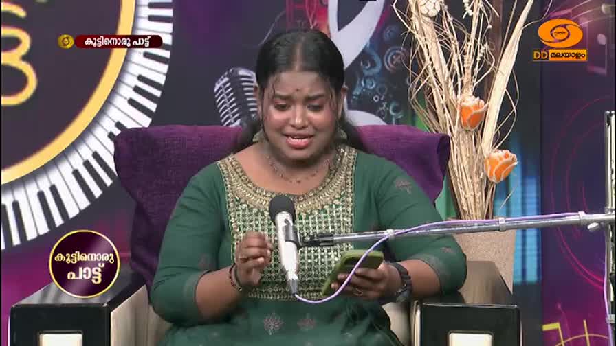 A young woman in a green dress sings into a microphone, her eyes closed. She holds a phone in her hand, looking down at it between verses.