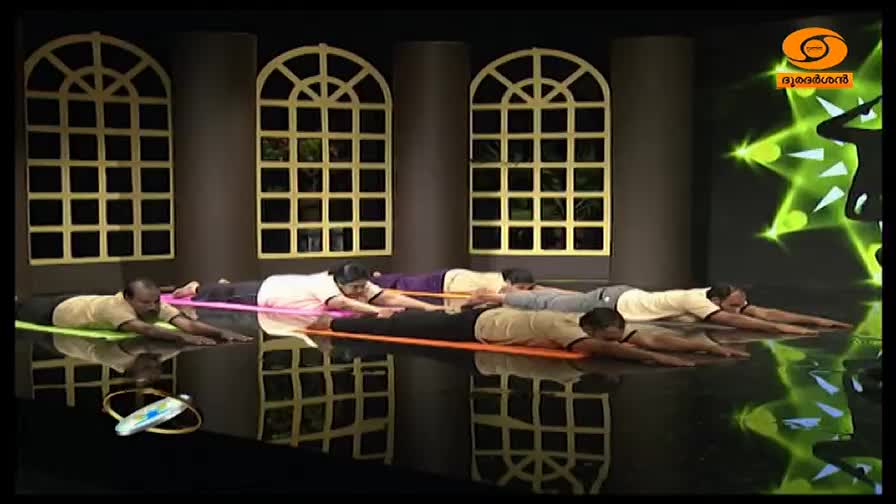 A group of people lies on yoga mats, their forearms extended forward. On the screen behind them, the DD Malayalam logo is visible.