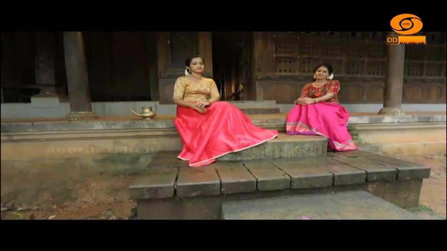 Two women in traditional Indian attire sit on stone steps outside a building. The woman on the left wears a gold blouse and a flowing pink skirt, while the woman on the right is dressed in a patterned red top and a bright pink skirt.