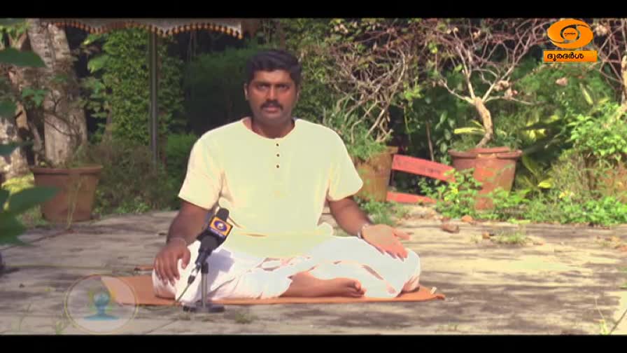 A man sits cross-legged on a mat in a garden, a microphone positioned before him. He wears a simple cream-colored kurta and white dhoti, his hands resting gently on his knees.