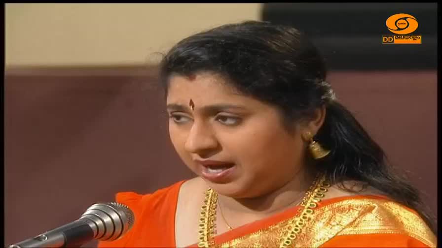 A woman in a bright orange sari, adorned with gold jewelry, speaks into a microphone. The DD Malayalam logo is visible in the corner of the screen.