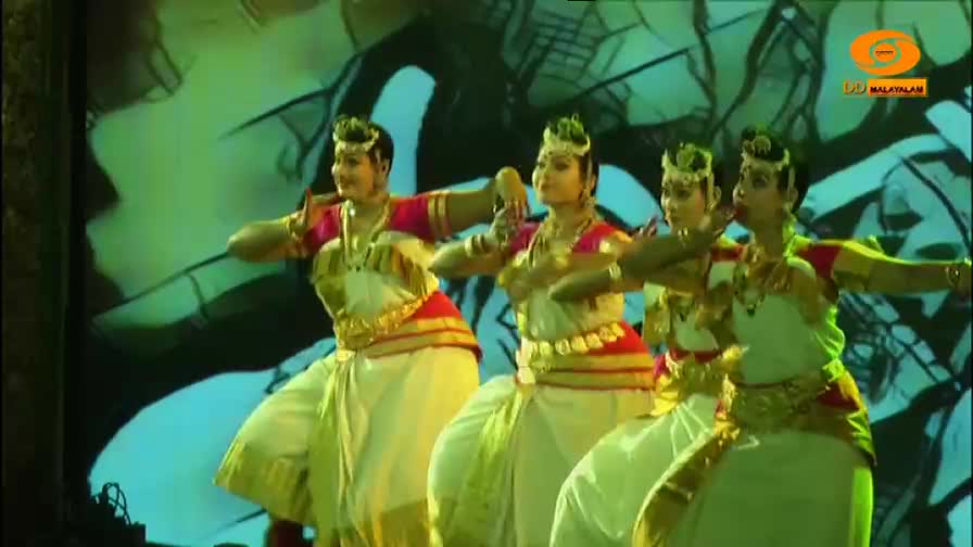 Four women in traditional Indian dance attire move in unison. Their hands are raised in expressive gestures against a backdrop of projected imagery.