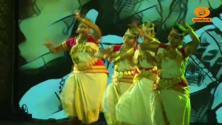 Four women in traditional attire perform a dance, their hands gracefully raised. The stage is lit with vibrant green and blue hues, a backdrop of abstract shapes behind them.