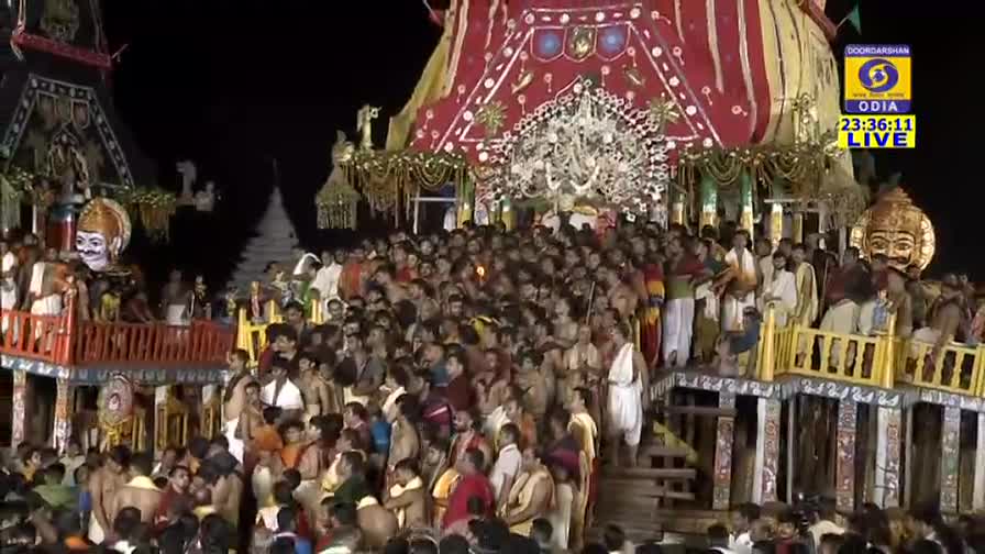 A massive crowd surrounds a brightly decorated chariot, its red and gold facade illuminated against the night. People are packed together, many reaching towards the towering structure as the live broadcast from DD Odia continues.
