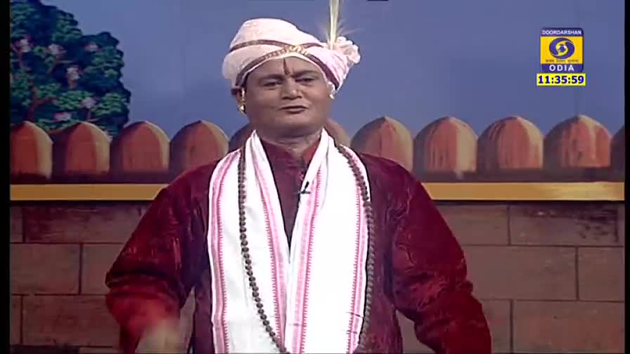 A man in a turban and traditional attire stands center stage, speaking into a small microphone. Behind him, a painted backdrop depicts a stylized wall, and the DD Odia logo is visible in the corner.
