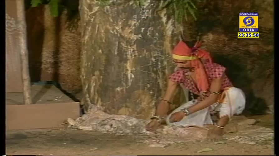 A man in traditional garb kneels, his hands working on a pile of white material. The scene is set against a backdrop of a textured, artificial tree, likely on a DD Odia production.
