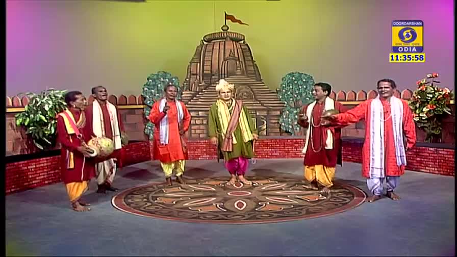 A group of costumed performers stand on a stage, with a painted temple backdrop behind them. One man in the center, wearing a turban and green robes, is the focal point, while others flank him, some holding instruments.
