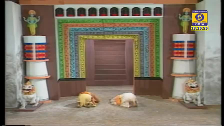 Two figures, dressed in white and yellow, are kneeling before a brightly painted doorway. The scene is set on DD Odia, with the time displayed in the corner.
