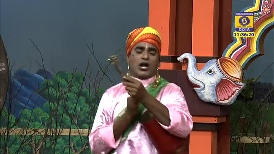 A man in a turban and pink shirt holds a small instrument, singing with his mouth wide open. The DD Odia logo and a painted elephant decorate the stage.
