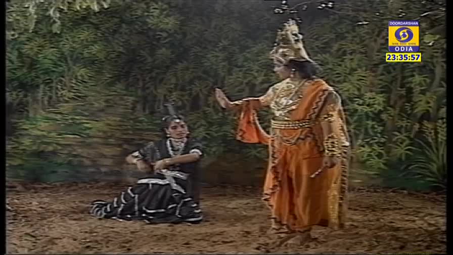 A figure in orange and gold gestures dramatically, while a woman in black and silver sits on the ground, her hands raised. The scene is set against a painted backdrop of trees, as if on Indian television.
