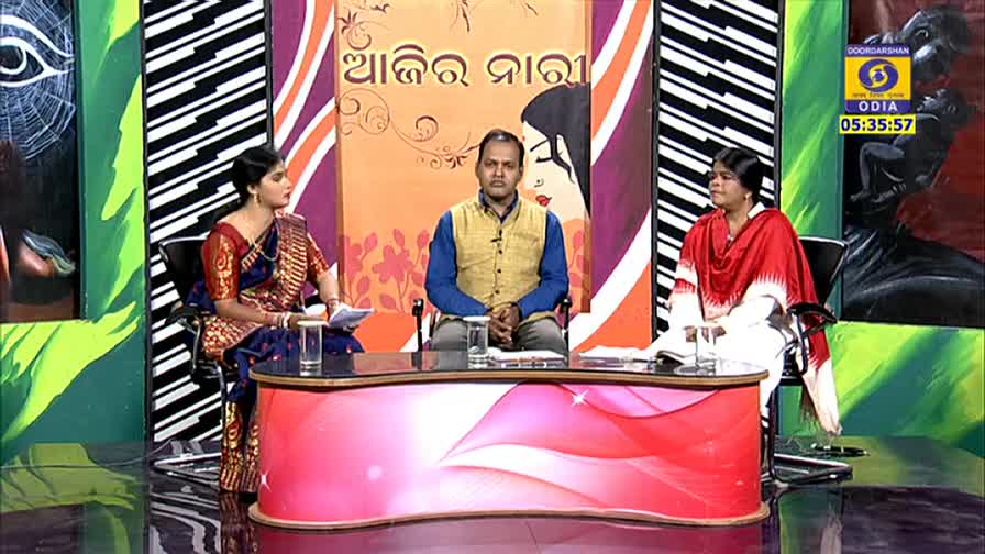 Three people sit behind a desk, facing the camera. The woman on the left, in a blue and red sari, holds a paper and looks to the side.

