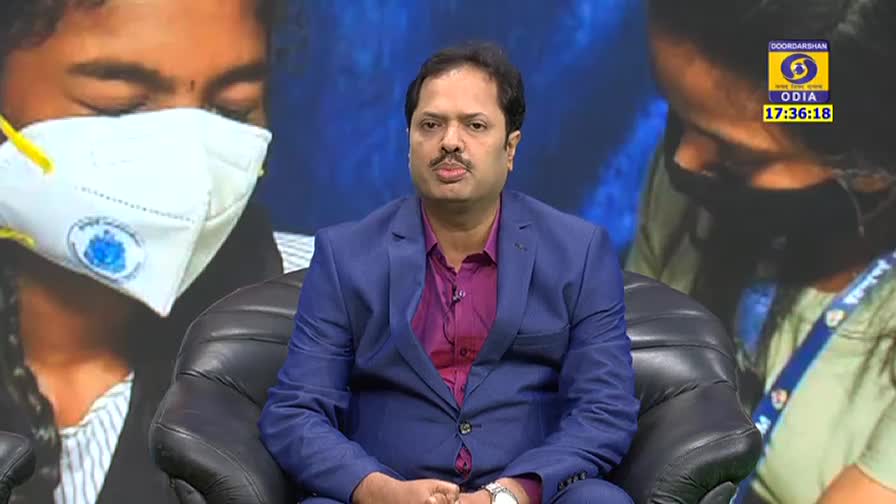 A man in a blue suit sits in a chair, speaking. Behind him, two figures are visible, one wearing a mask, and the DD Odia logo is in the corner.

