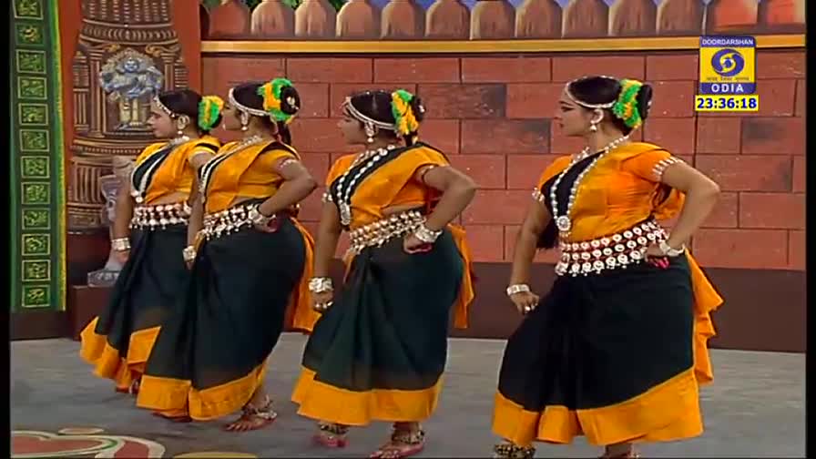 The dancers, in matching costumes of black and yellow, are in a line, their hands on their hips.  They're performing on a DD Odia set, the time displayed in the corner.
