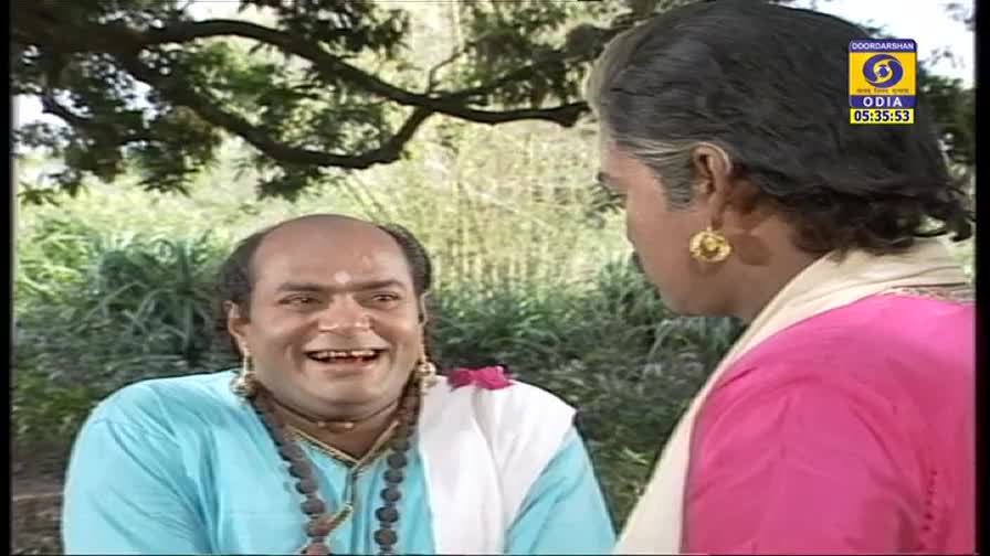 On DD Odia, one man in a light blue tunic is laughing, his mouth wide open, while another in pink looks on. The scene is set outdoors, under a tree, and appears to be a moment from a show.
