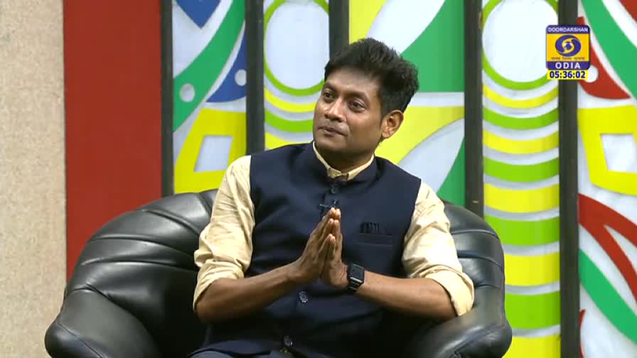 The man in the dark vest sits in a black chair, his hands clasped together in front of him. The DD Odia logo is visible in the corner, and the time is displayed.

