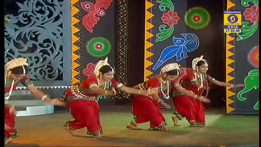 Dancers in red and white costumes are performing on a stage decorated with colorful painted designs. Their arms are extended, and their legs are in motion, as they move to the music on DD Odia.
