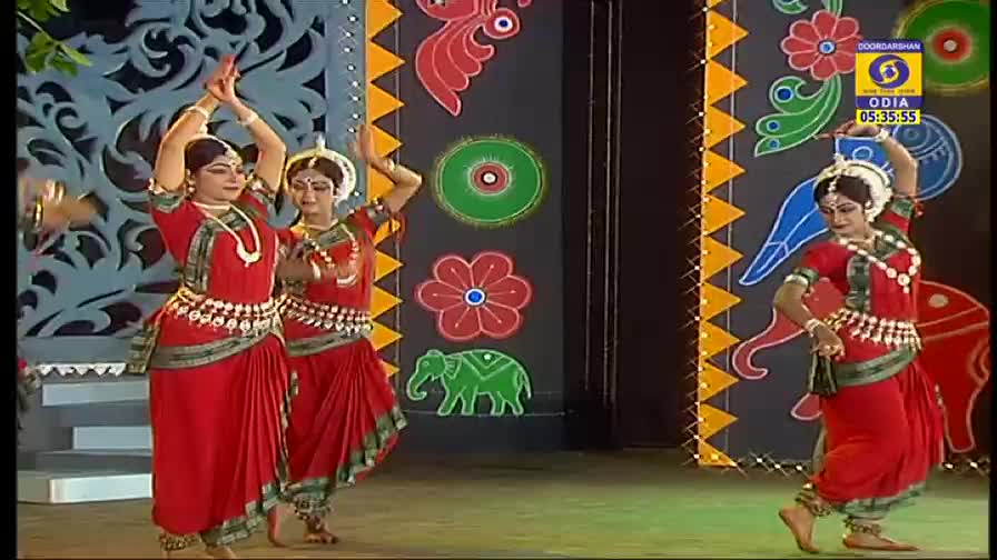 Dancers in vibrant red costumes gracefully move across the stage, their arms and hands forming elegant shapes. The performance, broadcast on DD Odia, is set against a backdrop of colorful, painted motifs.
