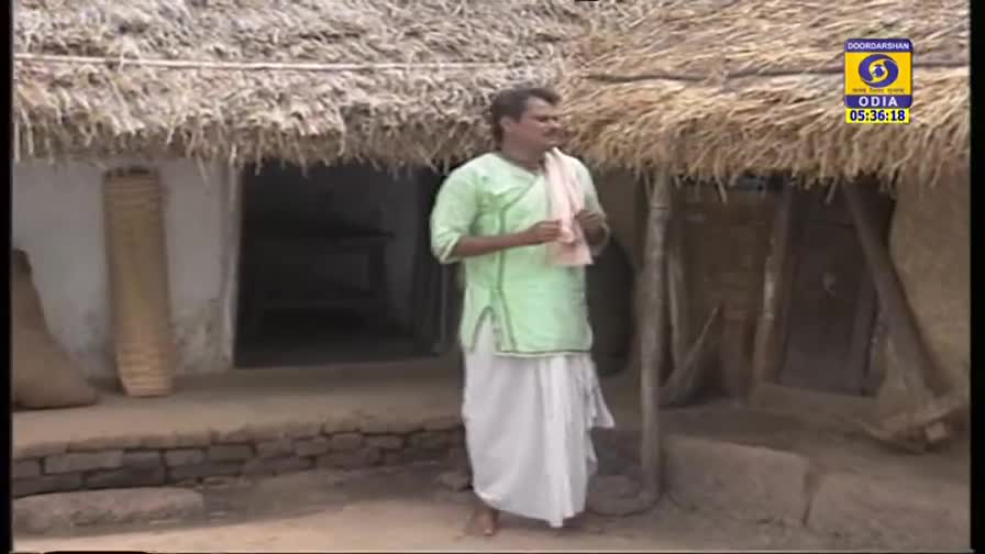 A man in a light green kurta and white dhoti stands in front of a thatched-roof house. The DD Odia logo in the corner indicates this scene is likely from a broadcast in India.
