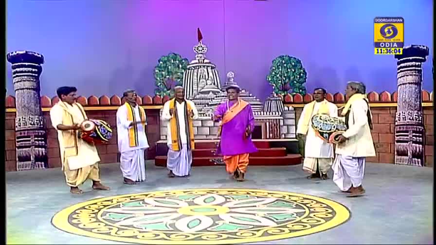 Six men in traditional dress are on a stage, some holding drums, while one in a purple outfit steps forward. A backdrop of a temple and a painted design on the floor complete the scene, likely for a performance on DD Odia.
