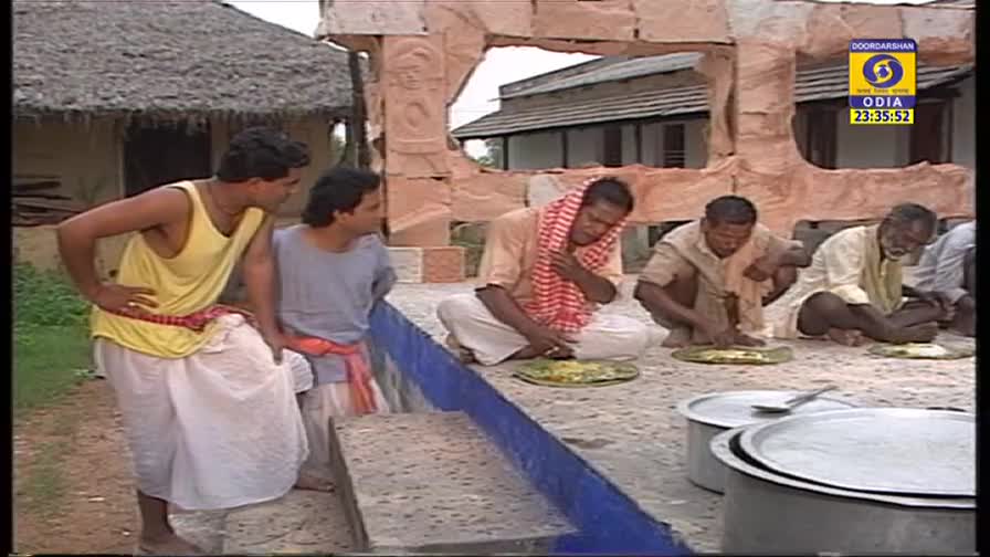 Men in traditional clothing sit on the ground, eating from plates. Two others, dressed in more modern attire, watch them from a nearby ledge.
