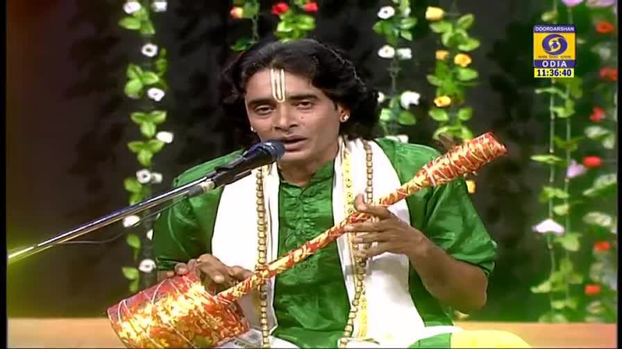 A man in a green shirt sings into a microphone, holding a long, decorated stringed instrument. The DD Odia logo is visible in the corner of the screen.

