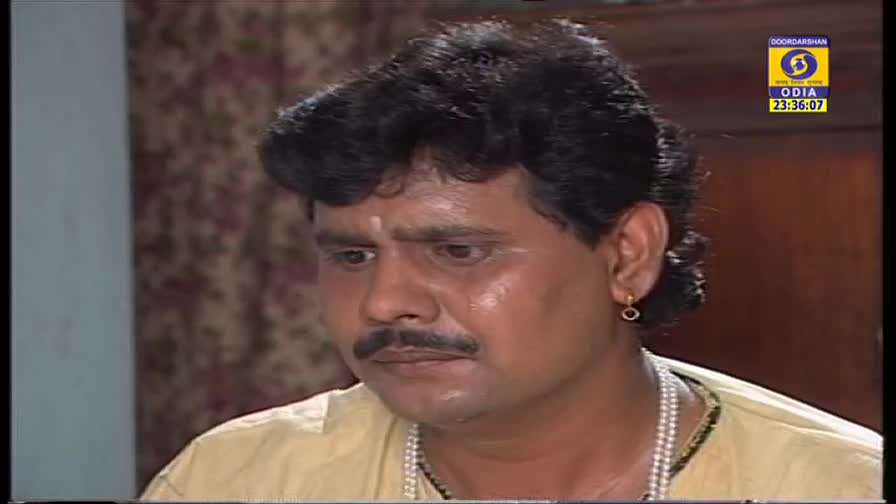 The man stares downward, his expression serious, as the DD Odia logo in the corner marks the time. He's wearing a cream-colored shirt and a necklace of pearls.
