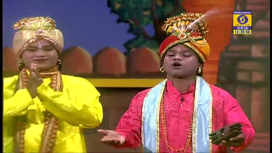 Two performers in ornate turbans and colorful robes are on stage, one gesturing with hands clasped, the other singing with a small instrument in hand. The DD Odia logo and the time are displayed in the corner of the screen.
