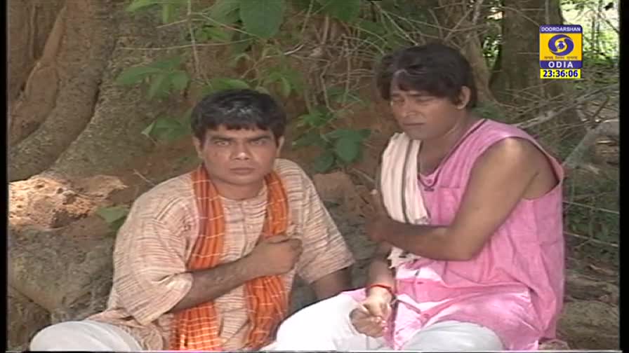 Two men sit near a large tree, one in a pink shirt and the other in a patterned shirt with an orange scarf. The DD Odia logo and time display in the corner.
