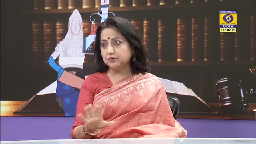 A woman in a red sari gestures with her hands as she speaks on a DD Odia program. The time is displayed on the screen, and a gavel sits on the table beside her.
